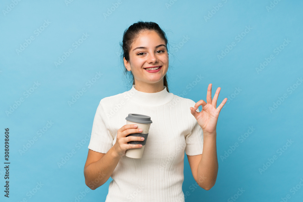 Young brunette girl over isolated blue background holding coffee to take away while making OK sign Young brunette girl over isolated blue background holding coffee to take away while making OK sign