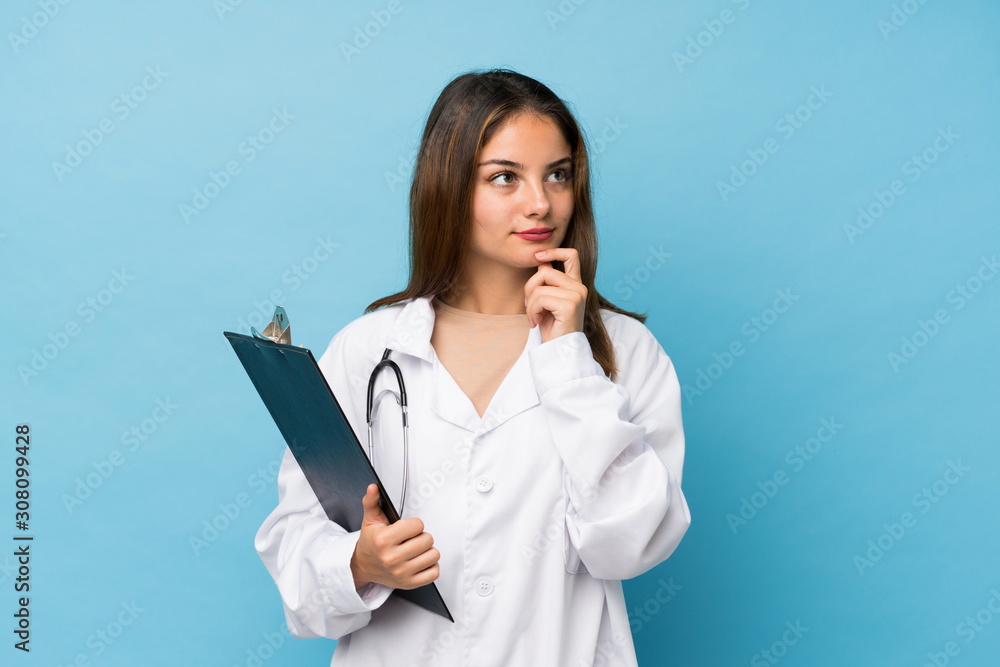 Young brunette girl over isolated blue background with doctor gown and holding a folder while thinking Young brunette girl over isolated blue background with doctor gown and holding a folder while thinking