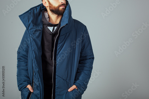 The guy model on a gray background is dressed in a blue down jacket. Hands in the pockets of a warm jacket. A guy with a beard and warmly dressed. Stylish warm clothes