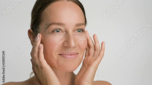 Close-up beauty portrait of young woman with smooth healthy skin, she gently touches her face with her fingers on white background and smiles