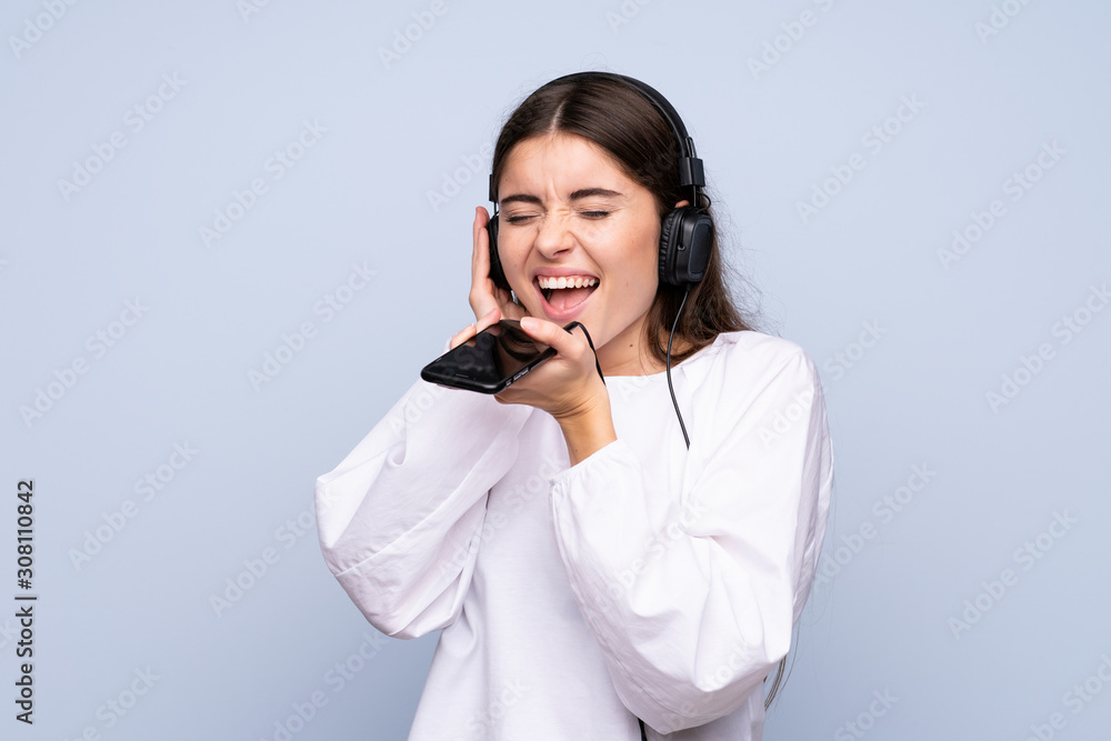 Young woman over isolated blue background using the mobile with headphones and singing Young woman over isolated blue background using the mobile with headphones and singing