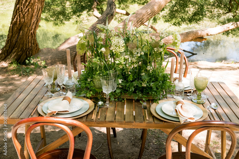 Festive table decor in openair next to the pond. Green color with wooden cutlery. Different natural white flowers. Wooden table and chairs. Wine glasses. Luxury wedding, party, birthday.