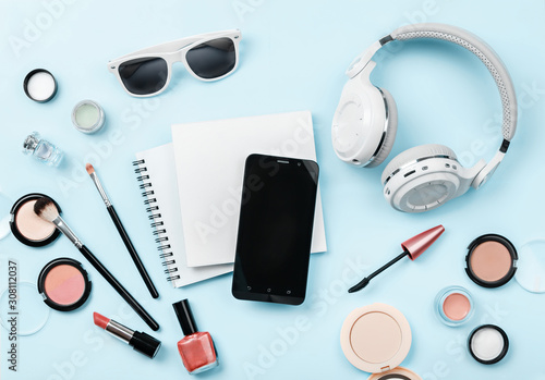 woman's cosmetics, phone, headphones and glasses on blue background. Notebook and decorative cosmetics: highlighter, concealer, rouge, and brushes for face make up. Copy space. 'To do' list Mock up