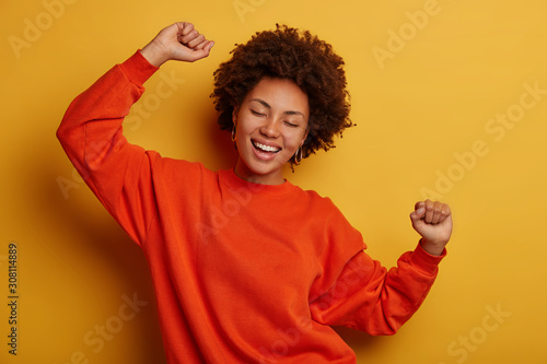 Tableau sur toile Photo of joyful brunette woman has fun and dances with hands raised, dressed in casual jumper, cheers over yellow background, gets promotion or approval, celebrates win