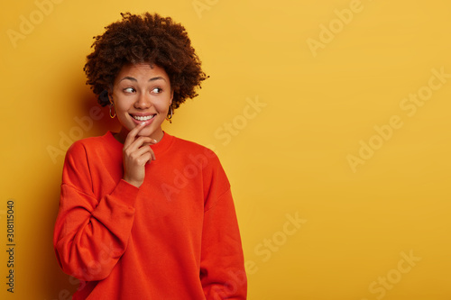 Charming tender curly woman smiles gladfully, looks aside, wears casual clothes, stands against yellow studio wall, free space for your advertising, satisfied with good promo, being amused and lucky © wayhome.studio 