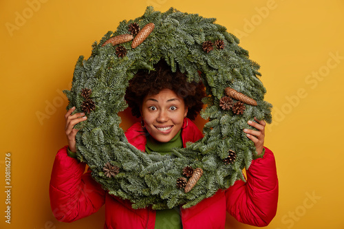 Close up shot of happy dark skinned woman looks through round spruce wreath, dressed in red jacket, makes shot against yellow wall, happy her favourite winter holidays come soon. Winter time photo