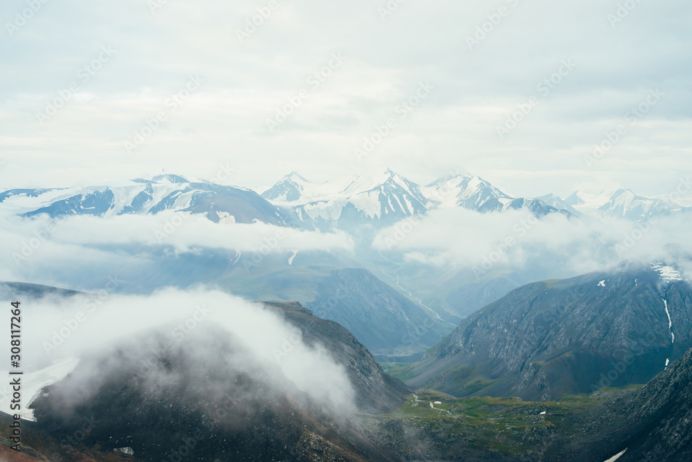 Beautiful atmospheric alpine landscape to big snowy mountains with glacier. Low clouds among rocks in green valley with small river. Wonderful highland scenery. Flying over mountains above clouds.