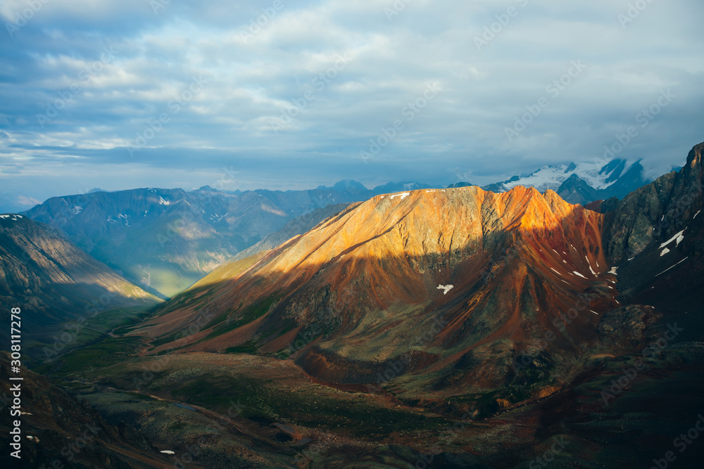 Atmospheric alpine landscape with red rockies in golden hour. Scenic green valley, big orange rocks and giant snowy mountains with glacier in sunrise. Wonderful highland scenery. Flying over mountains