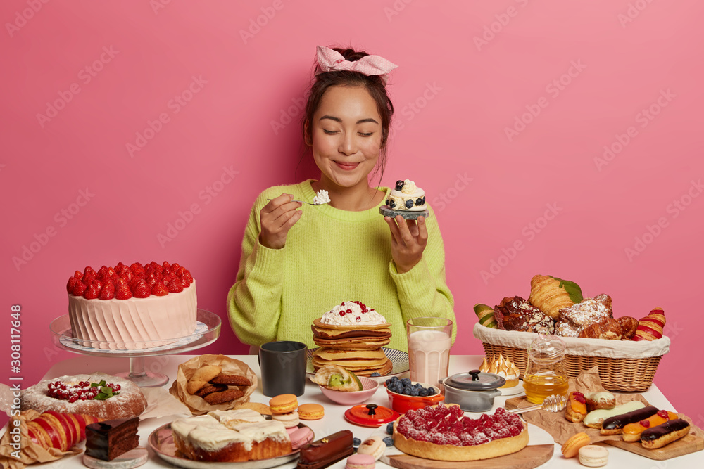 Photo of hungry sweettooth girl holds cupcake in one hand, spoon with cream in other, gets portion of sugar enjoys delicious snack during festive event keeps eyes closed from pleasure. Yummy treatment Photo of hungry sweettooth girl holds cupcake in one hand, spoon with cream in other, gets portion of sugar enjoys delicious snack during festive event keeps eyes closed from pleasure. Yummy treatment