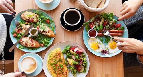 Different colorful meals for breakfast or lunch time on a plate with cutlery on woman's hands. Fried eggs, omelette, bruschetta and sausage on a wooden teble in restaurant. Flat lay top view panoramic photo