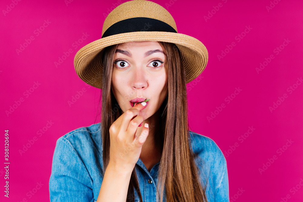 the rounded eyes of a young lady in a straw hat who chews strawberries speak of surprise. background pink