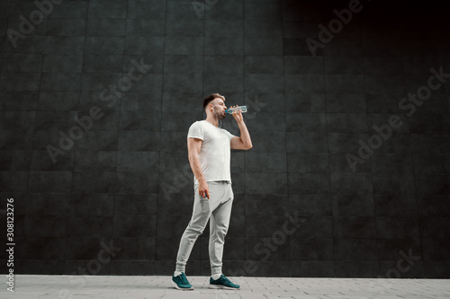 Fit handsome caucasian bearded blond sportsman drinking water and having earphones in ears while standing in front of dark background. © dusanpetkovic1