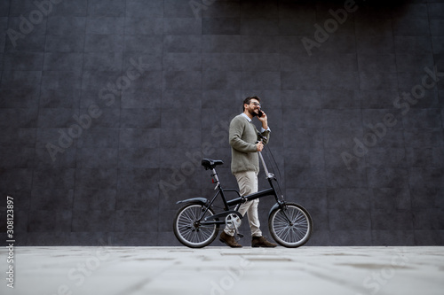 Side view of handsome caucasian fashionable man in sweater pushing bike and talking on the phone while passing by gray wall.