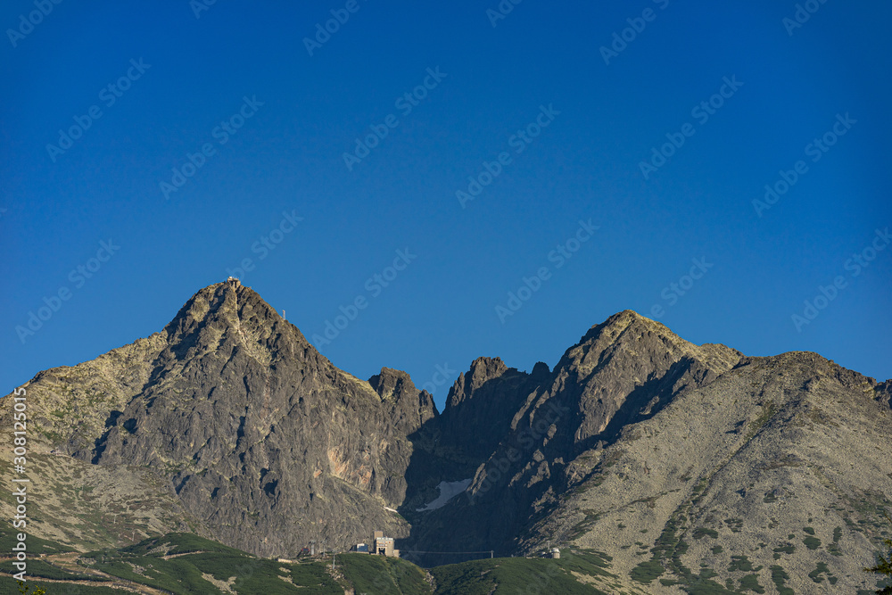 View of the Lomnicky stit (peak), famous rocky summit in High Tatras, Slovakia. Popular peaks Lomnicky stit and Kezmarsky stit on a sunny summer day with blue sky and no clouds. View of the Lomnicky stit (peak), famous rocky summit in High Tatras, Slovakia. Popular peaks Lomnicky stit and Kezmarsky stit on a sunny summer day with blue sky and no clouds.