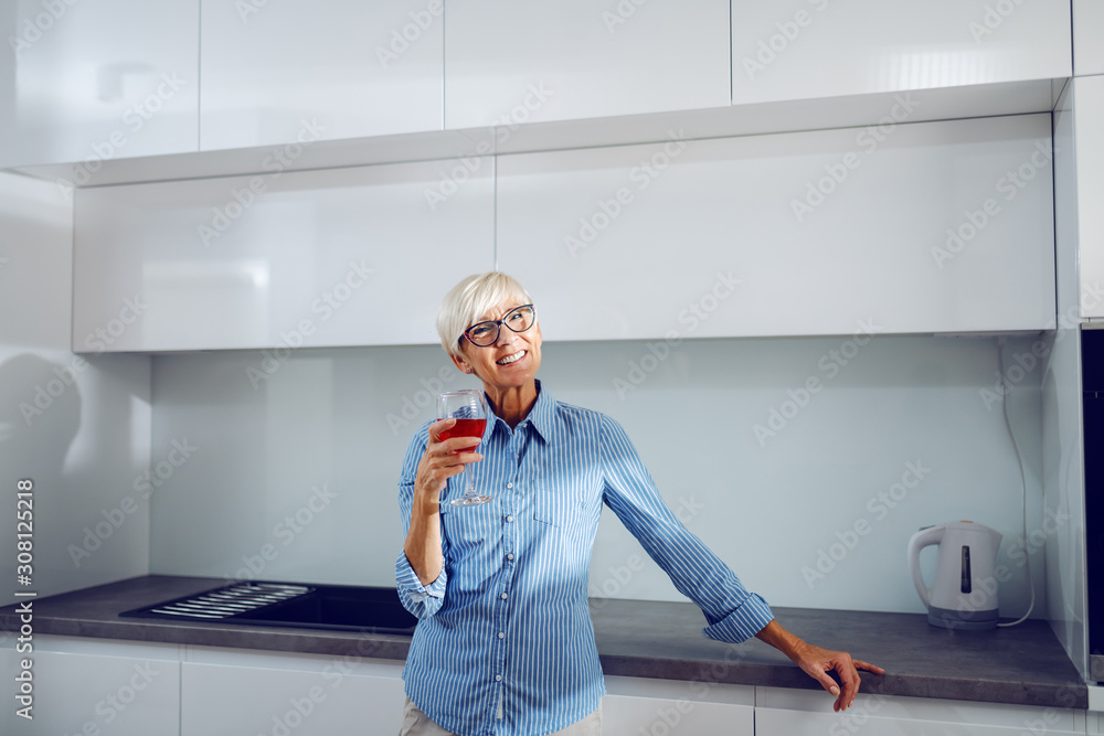 Charming smiling Caucasian senior blond woman leaning on kitchen counter and holding glass of red wine while looking at camera. Charming smiling Caucasian senior blond woman leaning on kitchen counter and holding glass of red wine while looking at camera.