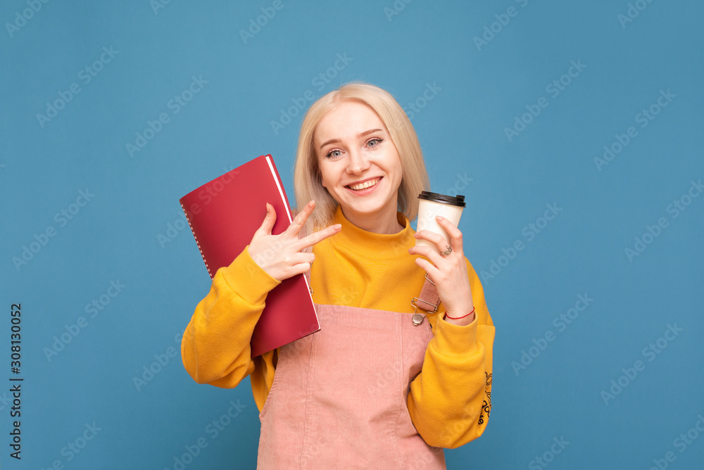 Portrait of a smiling student girl holds a cup of coffee and a notebook in her hands, wears a bright casual clothing, looks into the camera and smiles on a blue background. Isolated. Copyspace Portrait of a smiling student girl holds a cup of coffee and a notebook in her hands, wears a bright casual clothing, looks into the camera and smiles on a blue background. Isolated. Copyspace
