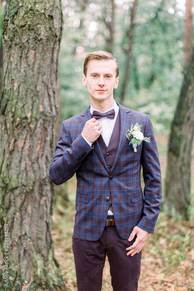 Trendy man groom in stylish suit touching his bow tie posing on the background of pine forest Trendy man groom in stylish suit touching his bow tie posing on the background of pine forest