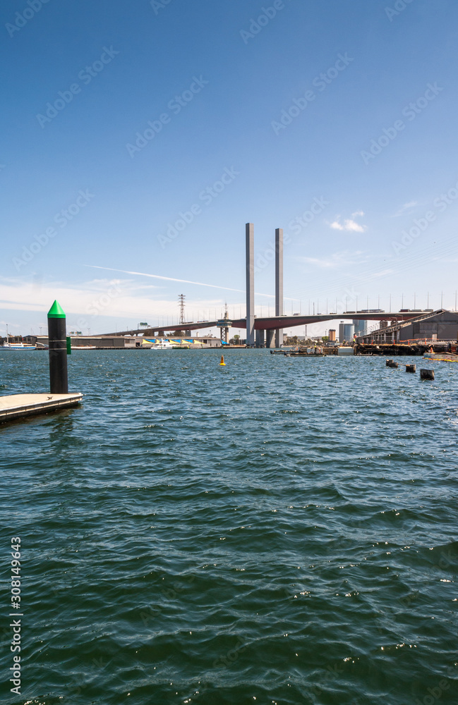 Melbourne, Australia - November 16, 2009: Portrait of Tall and large Bolte Bridge over Yarra River under blue cloudscape. High voltage line and tower. Green tip of short pier on blue water. Melbourne, Australia - November 16, 2009: Portrait of Tall and large Bolte Bridge over Yarra River under blue cloudscape. High voltage line and tower. Green tip of short pier on blue water.