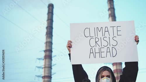 Young blonde woman putting up poster with slogan climate crisis ahead. Dirty air emissions of working factory on background. Polluted environment concept. Developing eco frienldy system. Industry photo