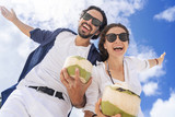 Beautiful young couple on a background of blue sky and clouds with coconuts in their hands. Joyful smiles and laughter. Honeymoon travel and vacation on the tropical ocean, picture from below