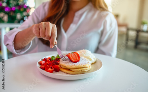 A beautiful asian woman holding and eating pancakes with strawberries and whipped cream by fork