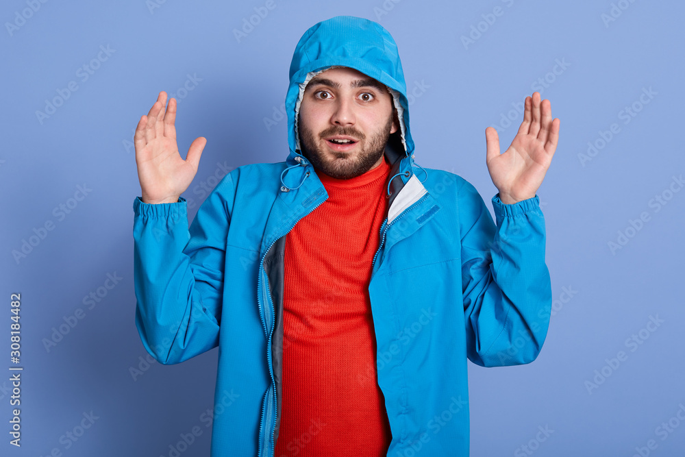Close up portrait of bearded man standing with hands up wearing blue jacket with hood and red shirt, posing isolated over studio background, looks scared, keeps mouth opened. People emotions concept.