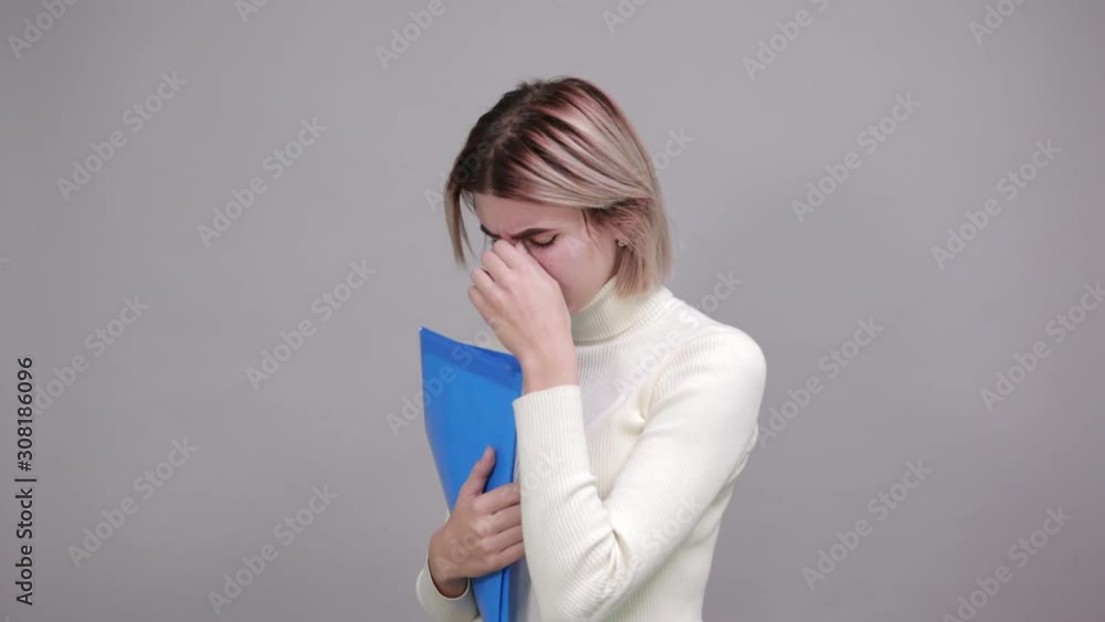 Strict young woman in white shirt, holds folder keeping hand on nose isolated on gray wall background in studio. Healthy fashion lifestyle, people sincere emotions concept. Strict young woman in white shirt, holds folder keeping hand on nose isolated on gray wall background in studio. Healthy fashion lifestyle, people sincere emotions concept.