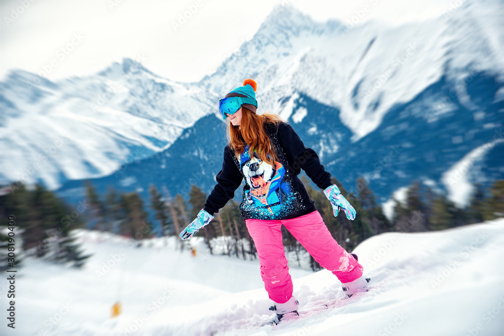 young happy girl rides on a snowboard in bright colored clothes on a background of snowy mountains