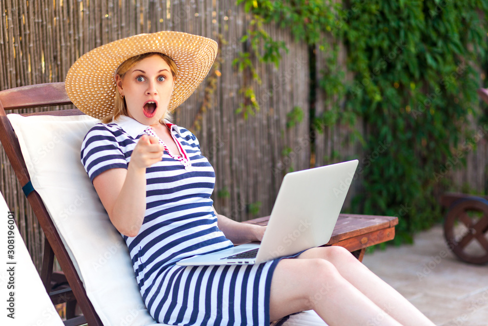 No way! Portrait of unbelievable young adult freelancer woman in hat and dress sitting on daybed with laptop, pointing finger and looking at camera with big eyes. Lifestyle concept, outdoor, summer,