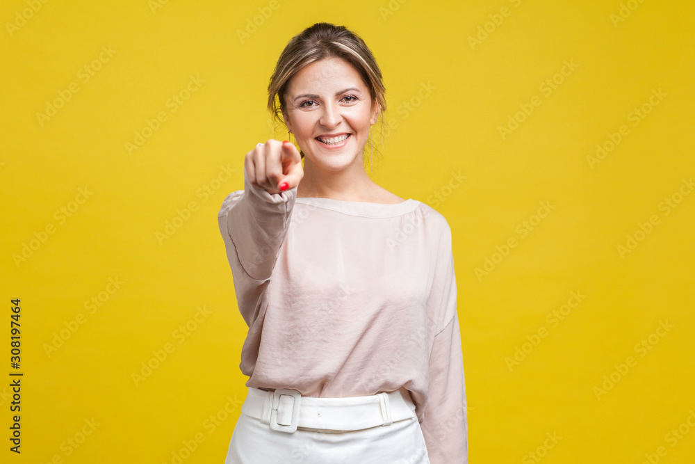 Portrait of cheerful young beautiful woman with blonde hair in casual beige blouse standing, looking at camera and pointing finger, successful choice, indoor studio shot isolated on yellow background Portrait of cheerful young beautiful woman with blonde hair in casual beige blouse standing, looking at camera and pointing finger, successful choice, indoor studio shot isolated on yellow background
