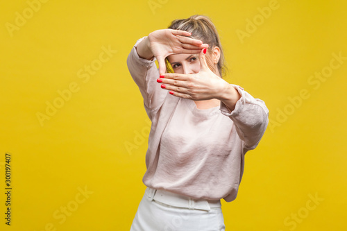 Fotografía Portrait of curious young woman with fair hair in casual beige blouse standing, making portrait or photo frame with hands, focusing looking at camera