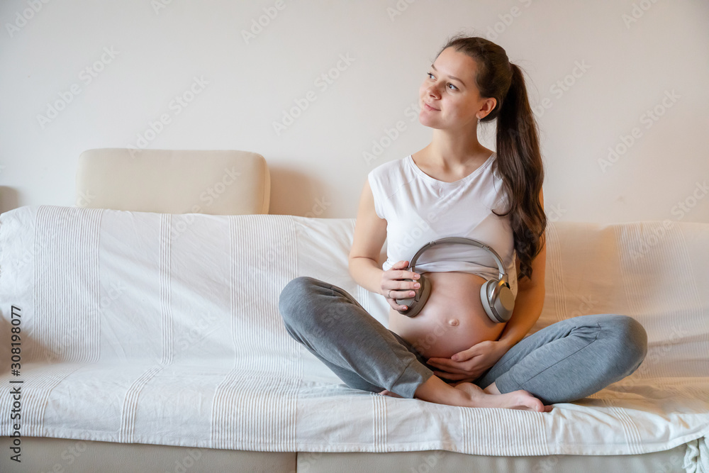 Pregnant woman sitting on white sofa at home and keeping headphones on her belly Pregnant woman sitting on white sofa at home and keeping headphones on her belly