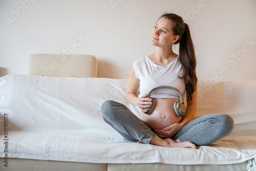 Pregnant woman sitting on white sofa at home and keeping headphones on her belly