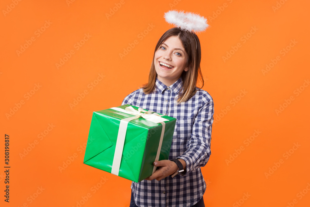 Portrait of lovely cheerful young woman wearing checkered shirt standing with halo on head, holding gift box, concept of generosity and charity. indoor studio shot isolated on orange background Portrait of lovely cheerful young woman wearing checkered shirt standing with halo on head, holding gift box, concept of generosity and charity. indoor studio shot isolated on orange background