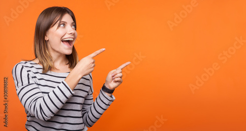 Portrait of surprised happy young woman with brown hair in striped shirt standing, pointing at empty space for announcement, unbelievable news. indoor studio shot isolated on orange background