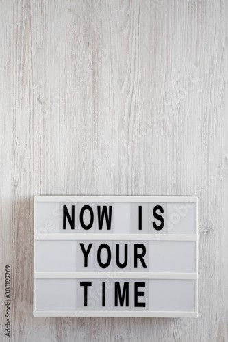 'Now is your time' words on a lightbox on a white wooden surface, top view. Overhead, from above, flat lay. Copy space.