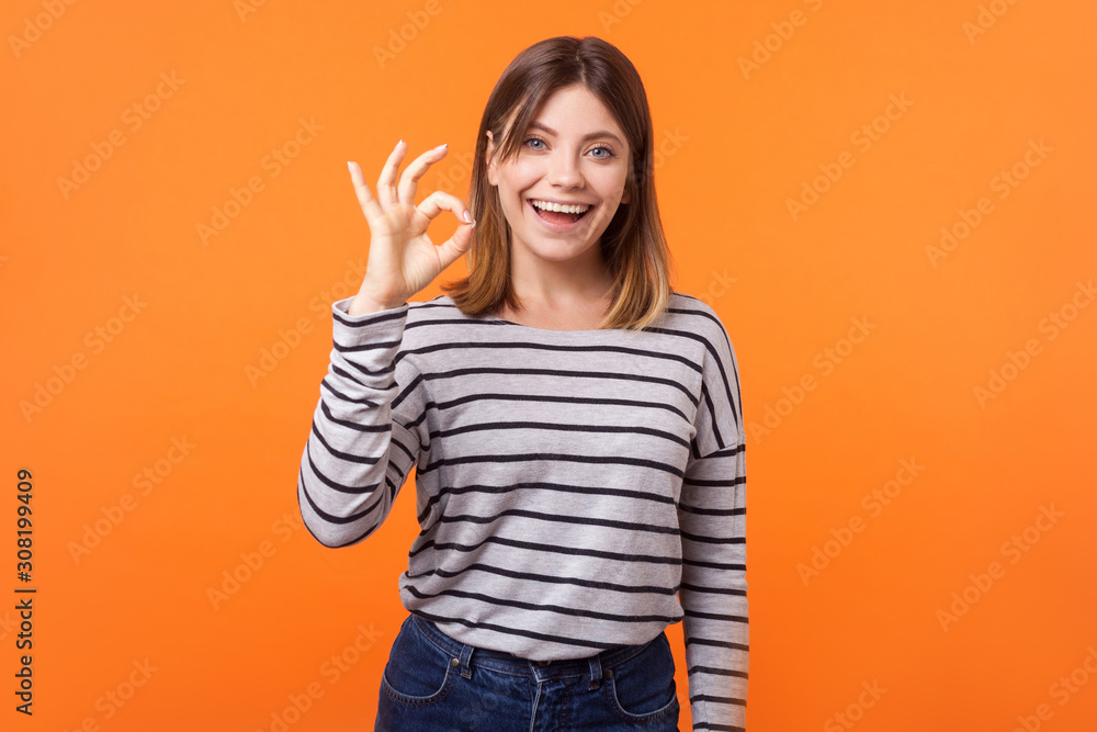 I am ok! Portrait of satisfied beautiful young woman with brown hair in long sleeve striped shirt standing showing okay gesture with fingers, good job. indoor studio shot isolated on orange background I am ok! Portrait of satisfied beautiful young woman with brown hair in long sleeve striped shirt standing showing okay gesture with fingers, good job. indoor studio shot isolated on orange background