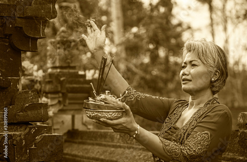 beautiful and happy Asian Indonesian woman dressed in traditional Balinese religious custom holding incense stick and flowers offering outdoors at Bali temple photo