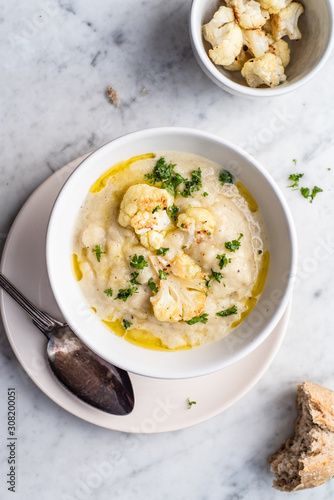Roasted cauliflower cream soup on white kitchen table, served with fresh parsley, olive oil and bread. Overhead view.