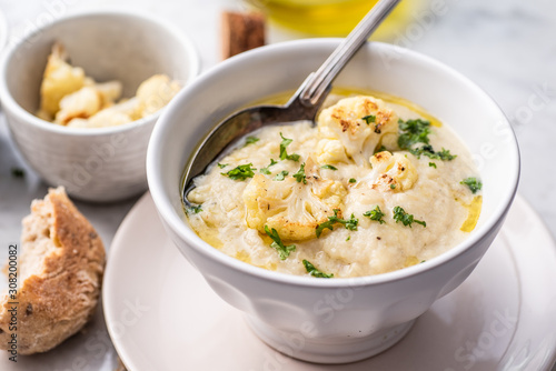 Roasted cauliflower cream soup on white kitchen table, served with fresh parsley, olive oil and bread.