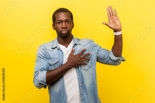 I swear! Portrait of honest serious man in denim casual shirt with rolled up sleeves keeping hand on chest and raising palm, giving promise, pledge. indoor studio shot isolated on yellow background