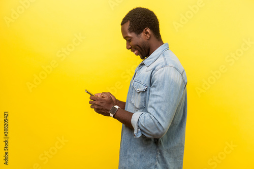 Quadro su tela Profile of positive smiley man in denim casual shirt using cellphone, typing text message or dialing number with joyous expression, technology concept