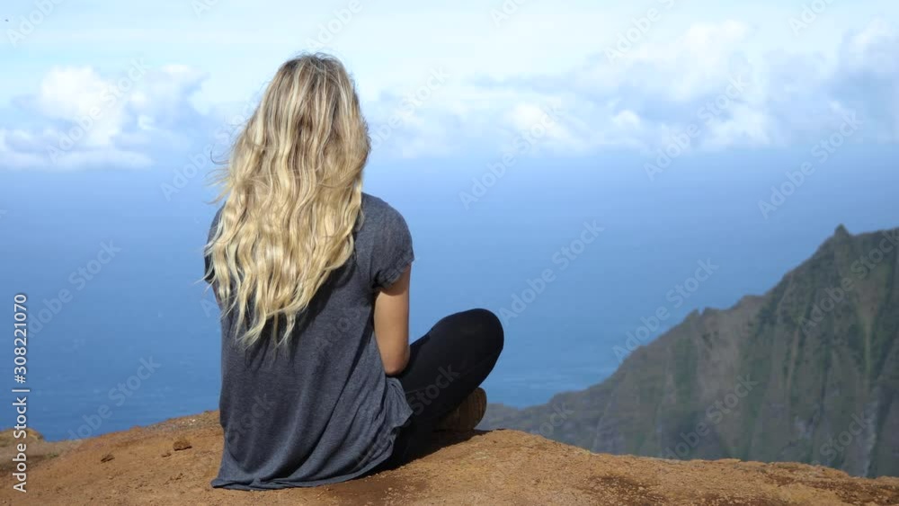 A blonde young woman sitting on cliff in the Napali Coast State Park in Kauai, Hawaii A blonde young woman sitting on cliff in the Napali Coast State Park in Kauai, Hawaii