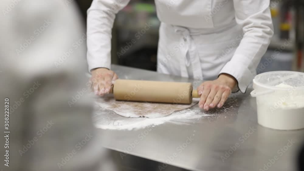confectioner in white uniform with a rolling pin rolls out the dough on a steel table sprinkled with flour confectioner in white uniform with a rolling pin rolls out the dough on a steel table sprinkled with flour