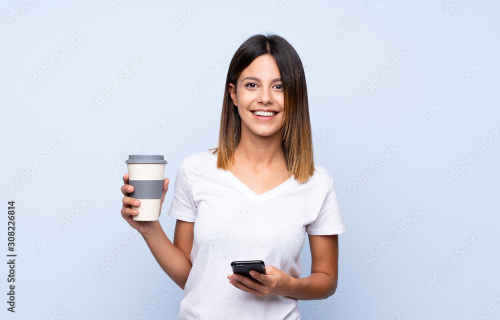 Young woman over isolated blue background holding coffee to take away and a mobile Young woman over isolated blue background holding coffee to take away and a mobile