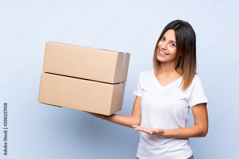 Young woman over isolated blue background holding a box to move it to another site Young woman over isolated blue background holding a box to move it to another site