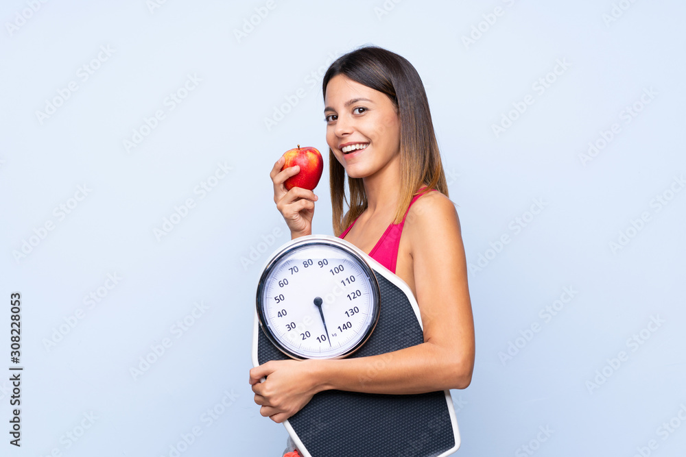 Girl with weighing machine over isolated blue background with weighing machine and with an apple Girl with weighing machine over isolated blue background with weighing machine and with an apple