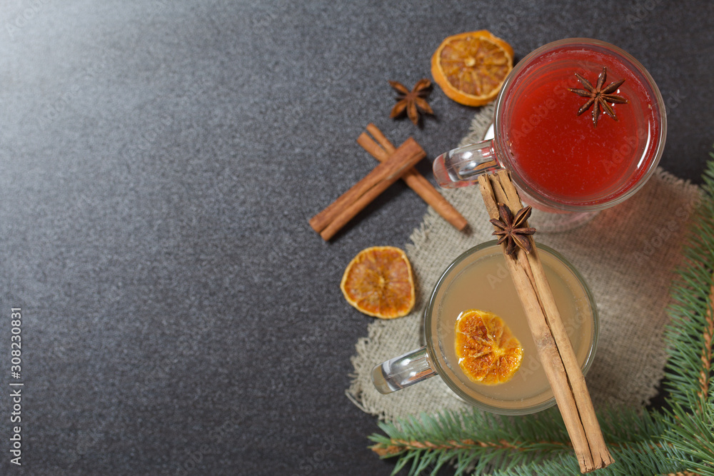 Glasses with a drink of orange and red are on a piece of linen. Slices of dried orange and anise float in them. Nearby are cinnamon sticks and dried oranges and a spruce branch.