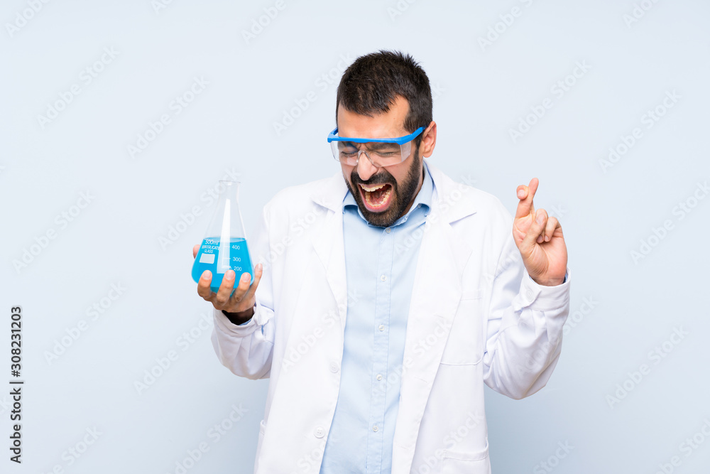 Young scientific holding laboratory flask over isolated background with fingers crossing