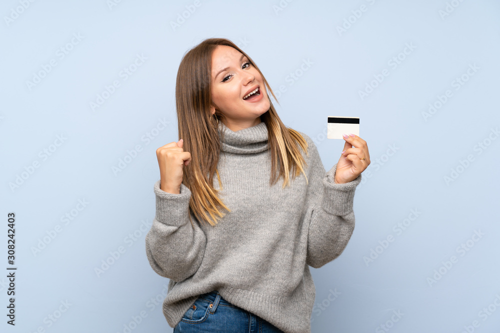 Teenager girl with sweater over isolated blue background holding a credit card Teenager girl with sweater over isolated blue background holding a credit card